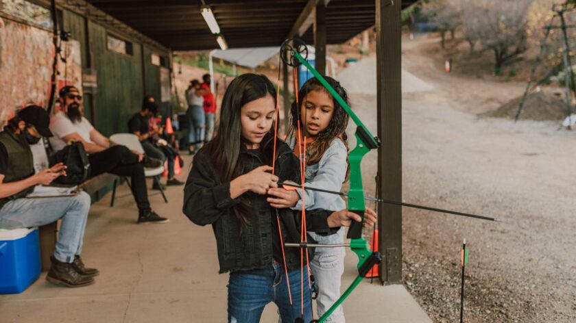 Two young girls practicing archery at an outdoor range, receiving guidance on bow handling.