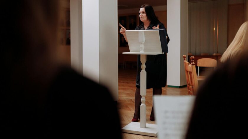 A woman conductor instructing a music class indoors, pointing and presenting.