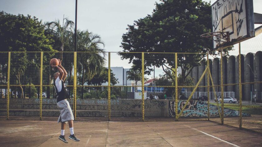 A man in athletic wear leaps for a basketball shot on an outdoor urban court.