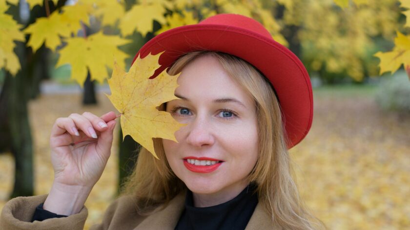 Smiling woman in red hat holding a fall leaf with vibrant autumn background.