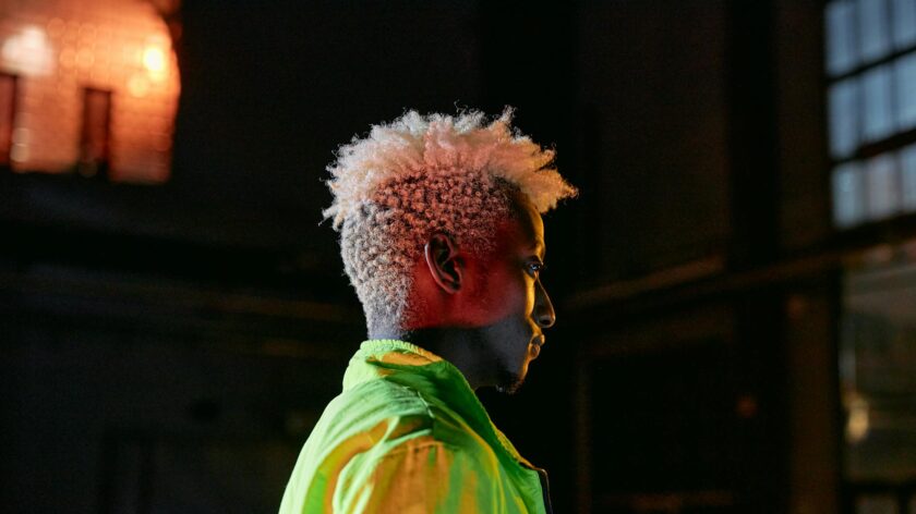 Side view of a man with afro hair wearing a neon green jacket, captured in dramatic lighting.