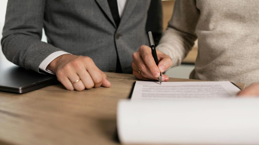 Close-up of businessmen signing documents at a wooden table in an office.