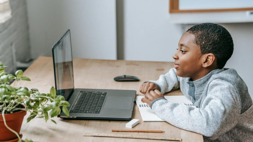 Side view of African American boy watching educational video with netbook while sitting at table with stationery